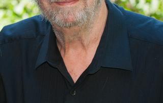 A white-haired man with a beard wearing a collared shirt smiles at the camera