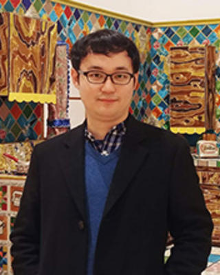 A young man with short black hair and glasses stands in front of a wildly colorful kitchen background, wearing a long black coat over a blue sweater and collared shirt.