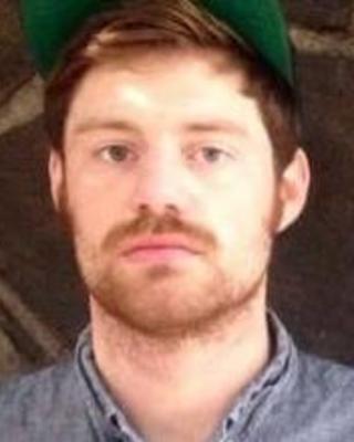 A man with short blonde hair and short facial hair wears a green baseball cap and looks at the camera with a serious expression