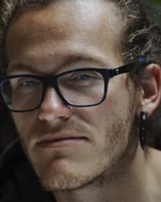 A nonbinary person with curly dark blond hair pulled back, wearing glasses, ear gauges, and a black T-shirt, looks pensively at the camera with an enigmatic expression