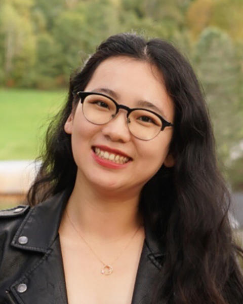 A woman with long black hair and glasses smiles in front of a scenic village
